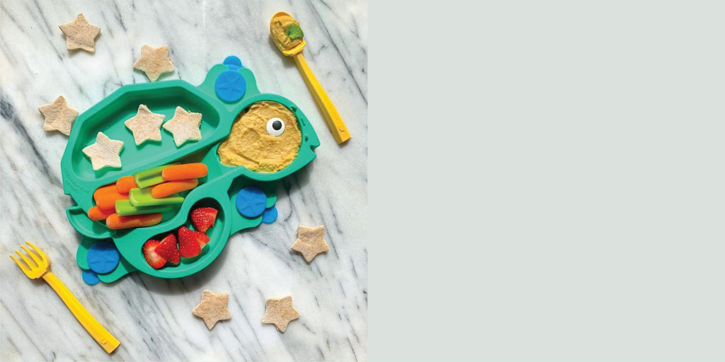 Image of the green Turtle Training Suction Plate styled with carrots, celery, hummus, and star shaped pita bread. Also featured are the yellow fork and spoon.