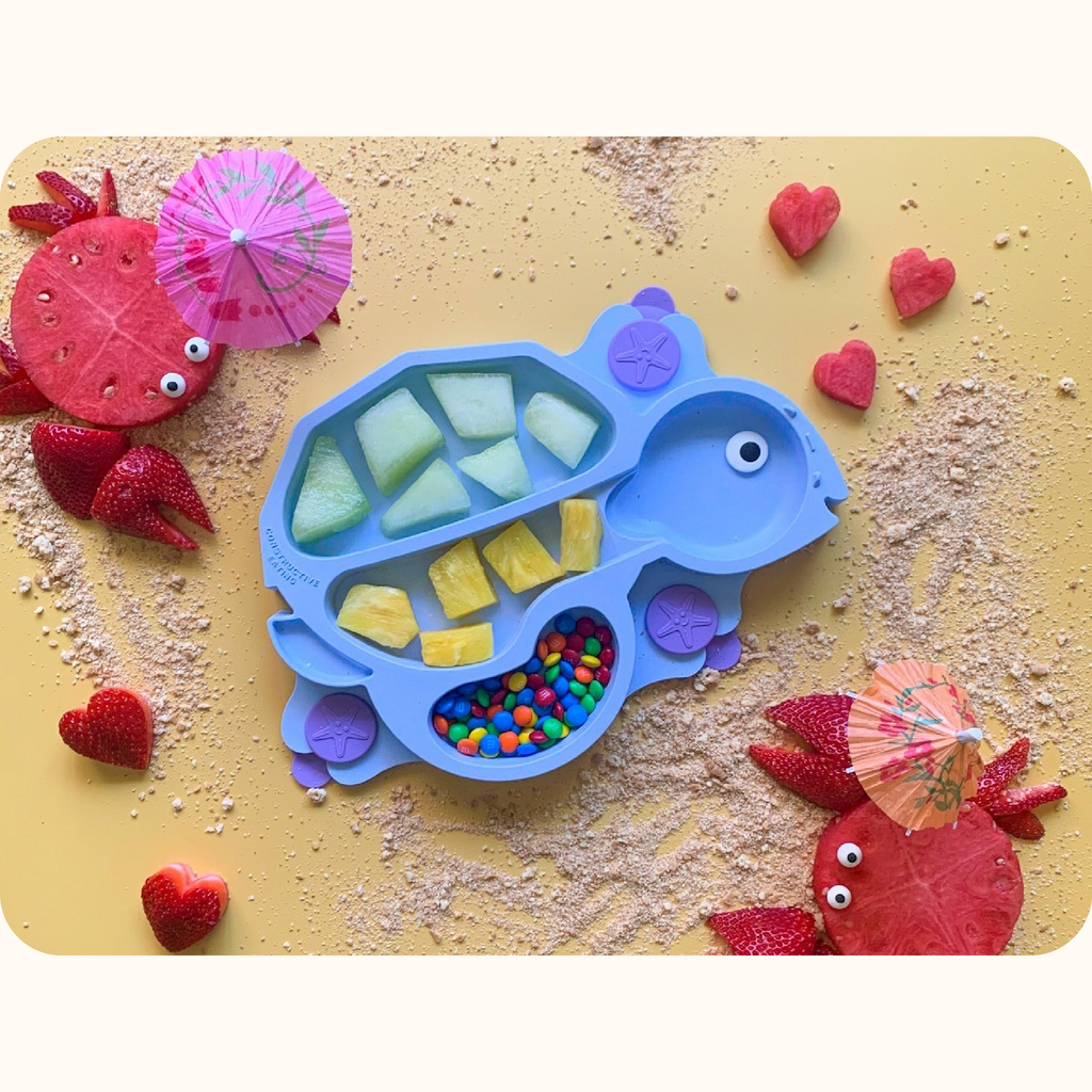 Image includes the blue turtle training suction plate. The plate is styled with honeydew, pineapple, and M&M's. To the sides of the plate there are crabs made out of watermelon and strawberries holding drink umbrellas. There are also heart-shaped strawberries and watermelon, and graham cracker crumbs.