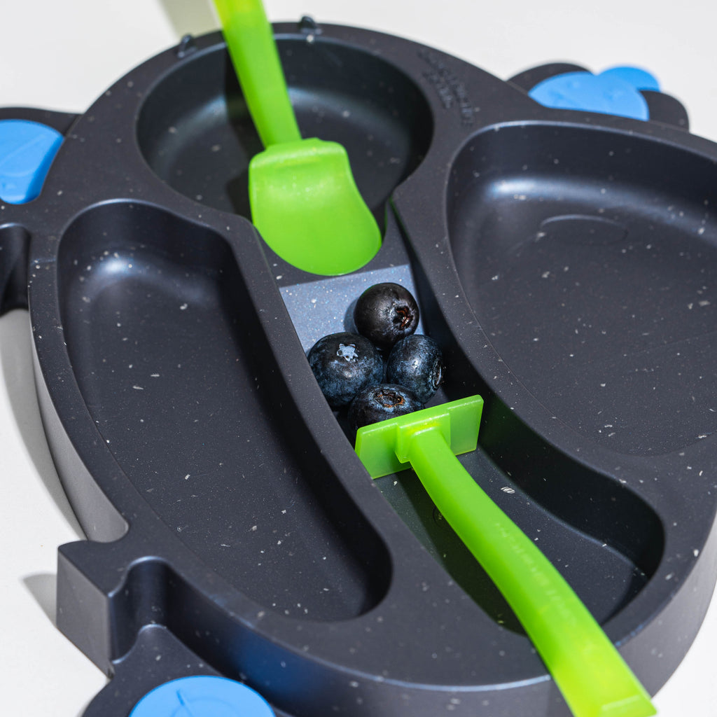 Image of the spacedust UFO Training Plate with the green pusher pushing blueberries up the patented ramp onto the green spoon. The green spoon is located in the spoon nest.