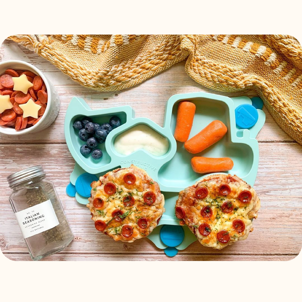 The image includes the teal truck training suction plate. The plate is styled with mini pizza bagels, blueberries, carrots, and ranch dressing. To the side of the plate there is a small bowl with mini pepperonis and star shaped cheese, and a seasoning shaker. Also pictured is a yellow blanket above the plate.