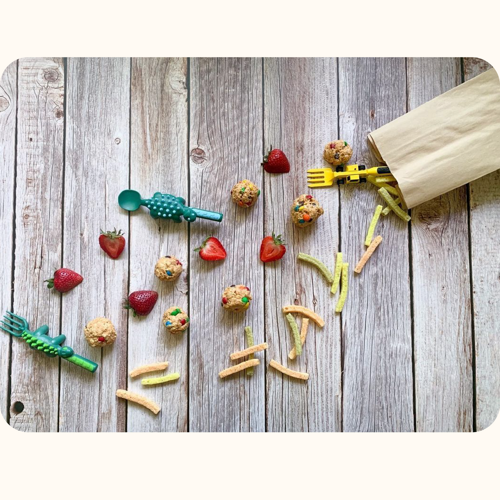 The image includes a yellow construction fork lift fork, a stegosaurus dino fork, and an ankylosaurus dino spoon. The utensils are coming out of a brown paper bag and in line with strawberries, veggie straws, and no bake oatmeal snack balls. 