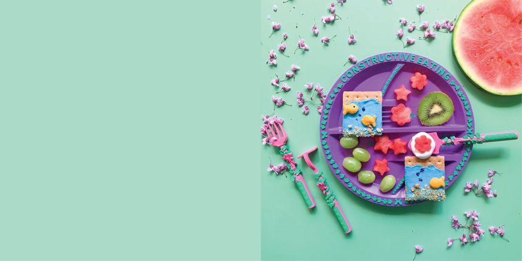 Image of the pink Garden Fairy Utensils and purple Plate styled with grapes, kiwi, watermelon in the shape of stars and flowers, and graham crackers. The graham crackers are styled with food to create an under the sea portrait. The food on the graham crackers include blue greek yogurt, goldfish, and graham cracker crumbs. The utensils included are a garden hoe pusher, a garden rake fork, and a garden shovel spoon.