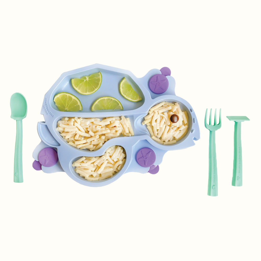 Image of the blue Turtle Training Plate styled with limes and plain macaroni noodles. Also pictured are the seafoam green training utensils which includes a pusher, fork, and spoon.