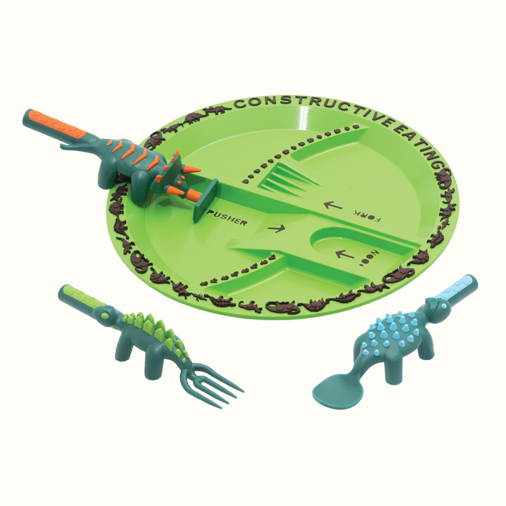 Image of the Dino Utensils and Dino Plate. The Dino Pusher is in the shape of a triceratops, the Dino Fork is in the shape of a stegosaurus, and the Dino Spoon is in the shape of an ankylosaurus.. The plate is orange. The pusher utensils is in its designated spot on the plate. The fork and spoon are angled in front of the plate.