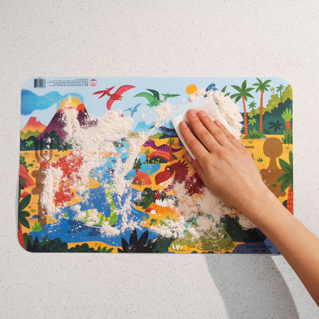Image of the Dino Placemat covered in flour with someone using a paper towel to wipe the surface clean.