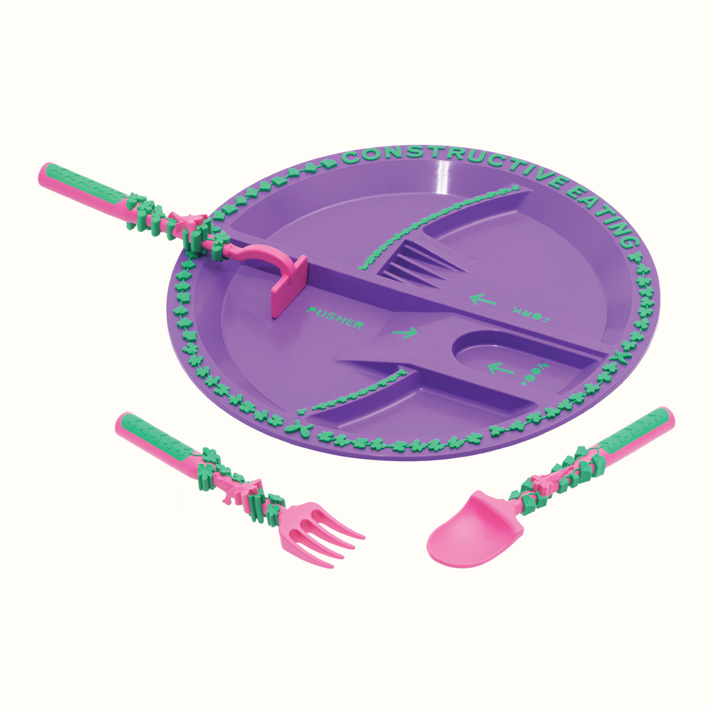 Image of the Garden Fairy Utensils and Plate. The pink utensils include a garden hoe pusher, a garden rake fork, and a garden shovel spoon. The plate is purple. The pusher utensil is in its designated spot on the plate. The fork and spoon are angled in front of the plate.