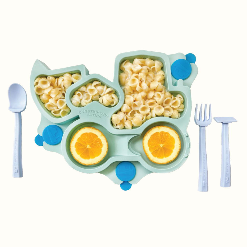 Image of the teal Truck Training Plate styled with oranges and plain shell noodles. Also pictured are the blue training utensils which includes a pusher, fork, and spoon.