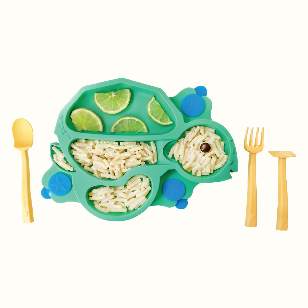 Image of the green Turtle Training Plate styled with limes and plain macaroni noodles. Also pictured are the yellow training utensils which includes a pusher, fork, and spoon.
