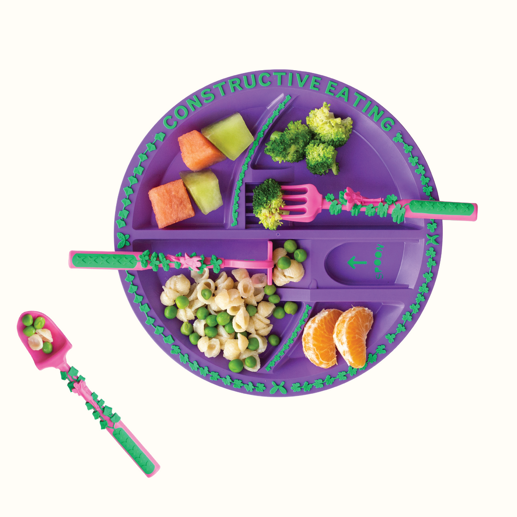 Image of the Garden Fairy Utensils and Plate styled with food. The pink utensils include a garden hoe pusher, a garden rake fork, and a garden shovel spoon. The plate is purple and is plated with honeydew, watermelon, broccoli, clementines, and a simple pasta with shell noodles and peas.