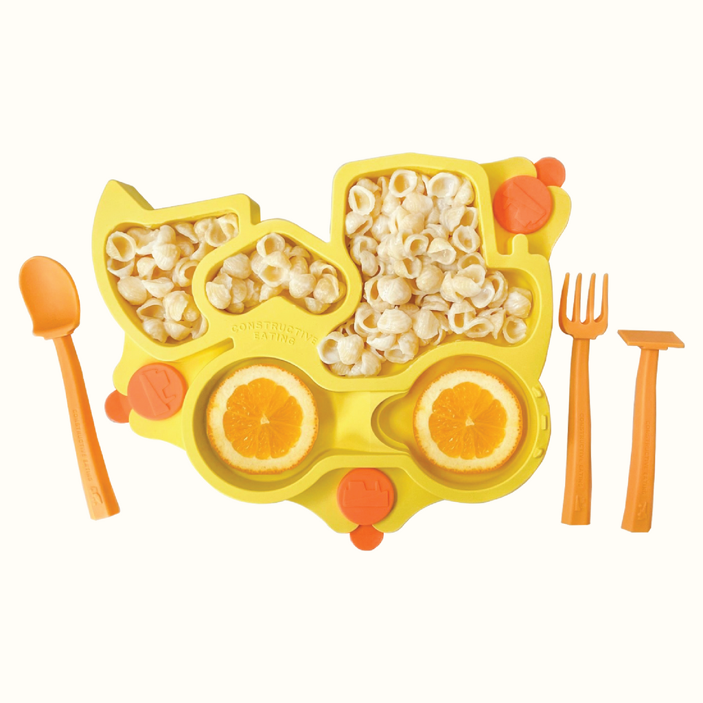 Image of the yellow Truck Training Plate styled with oranges and plain shell noodles. Also pictured are the orange training utensils which includes a pusher, fork, and spoon.